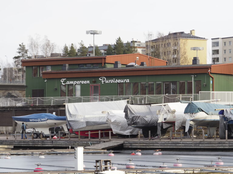 Kuvassa on vihreä Tampereen Pursiseuran -seurarakennus. Rakennuksen edessä on talvipeitteisiä pukeilla olevia purjeveneitä.